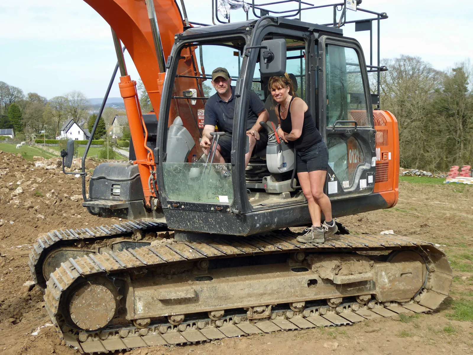 Dave and Louise Thompson standing beside freshly dug ground at the start of groundwork for the new covered arena, smiling and holding a shovel, with a rural farm setting behind them and a small wooden sign visible in the background reading New Arena Groundwork Underway