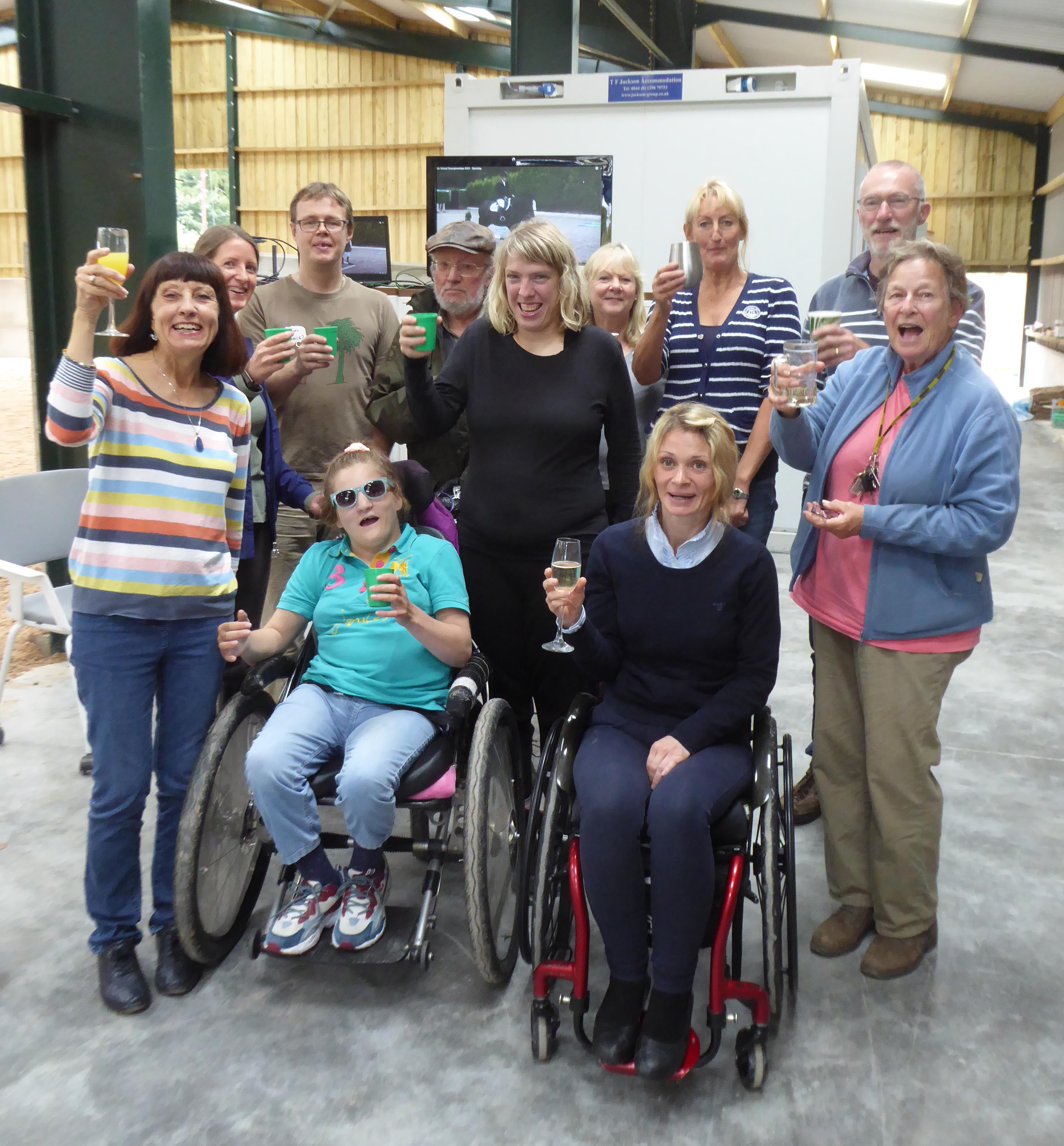 A group of riders, volunteers and supporters gather indoors at Buxton Riding School to celebrate Niamh Simmons after her success in the RDA Virtual National Championships 2021. They stand together smiling and raising glasses in a warm, joyful atmosphere. The room is a stable interior with a horse in the background and a large screen showing the competition results. Text visible on the screen reads RDA Virtual National Championships 2021 and Niamh Simmons.