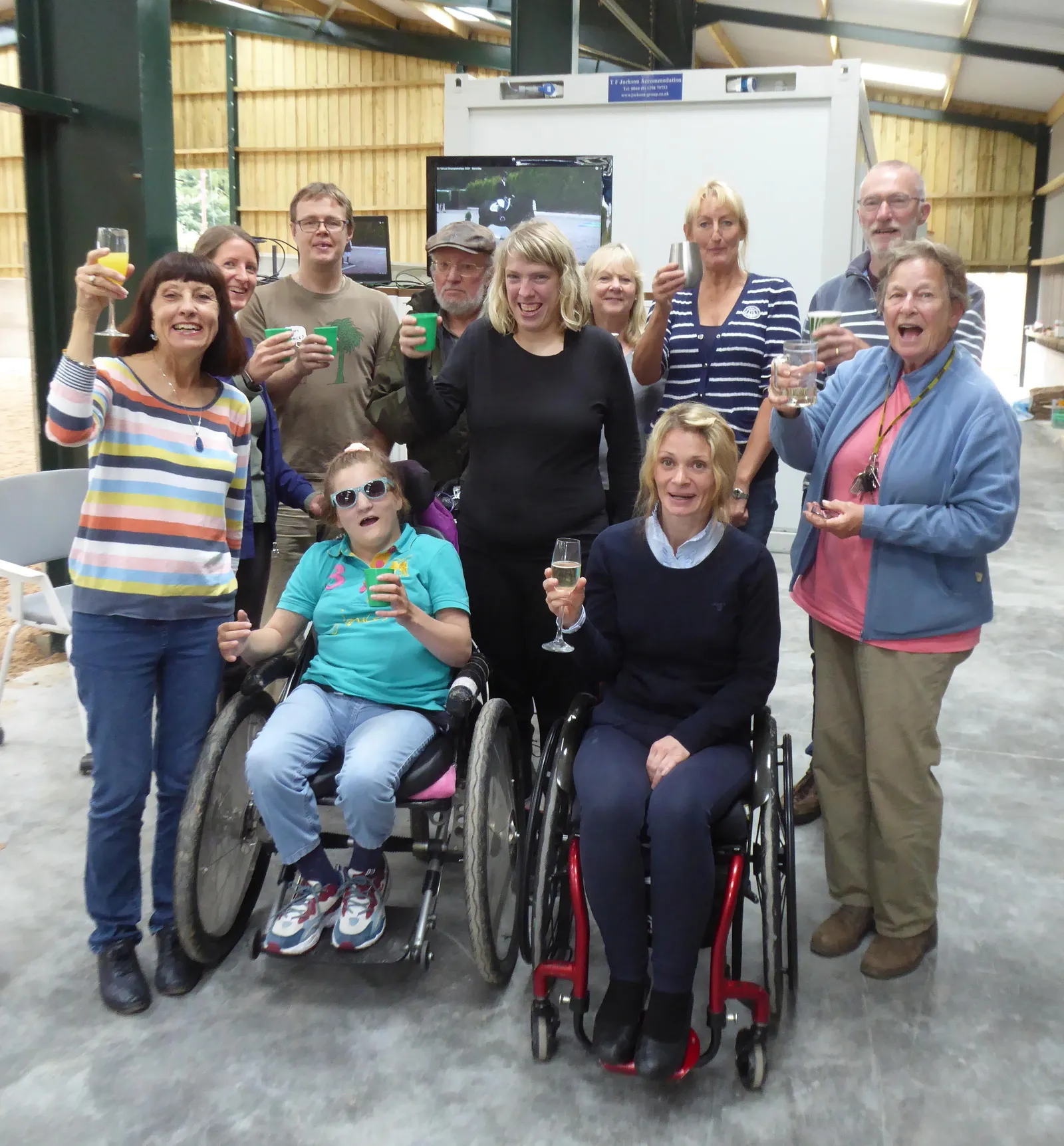 A group of riders, volunteers and supporters gather indoors at Buxton Riding School to celebrate Niamh Simmons after her success in the RDA Virtual National Championships 2021. They stand together smiling and raising glasses in a warm, joyful atmosphere. The room is a stable interior with a horse in the background and a large screen showing the competition results. Text visible on the screen reads RDA Virtual National Championships 2021 and Niamh Simmons.